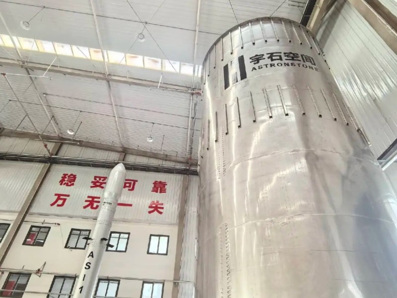 Astronstone's stainless steel rocket stage and AS-1 scale model inside a production facility in China, featuring prominent weld lines and branding; a large banner on the wall displays Chinese characters.