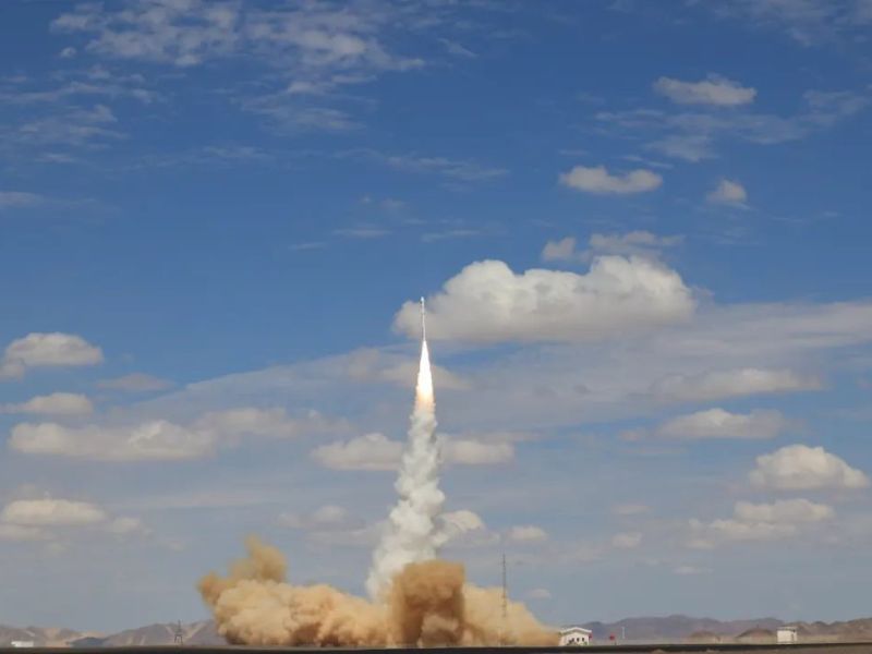 A 20-meter-tall rocket lifts off into the sky from a cloud of dust at the desert spaceport of Jiuquan, northwest China, with a trail of exhaust behind the rocket. The rocket is rising into a blue sky punctuated by a number of cumulus clouds.