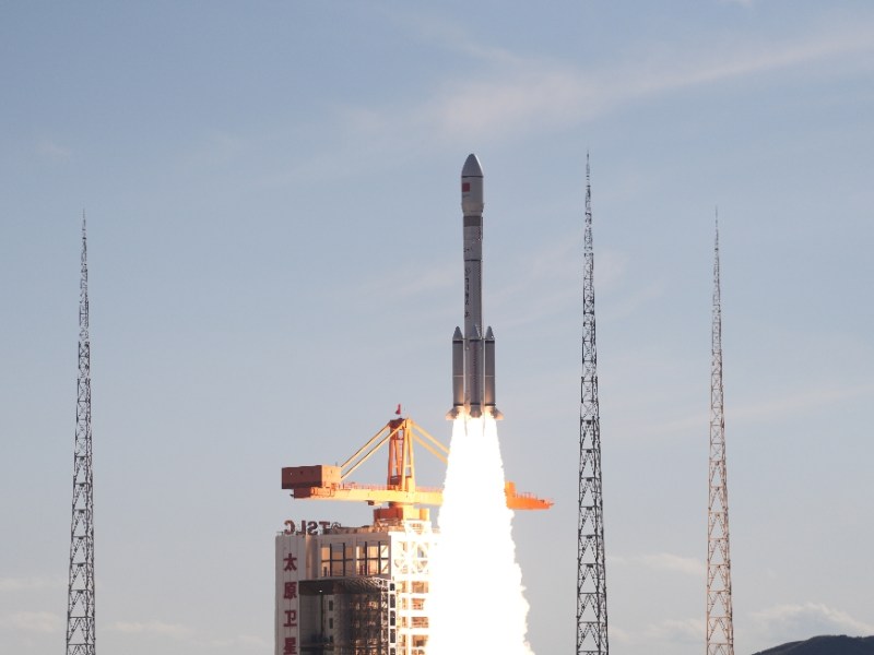 A Long March 6A rocket lifts off from the Taiyuan Satellite Launch Center in northern China, producing a bright plume of exhaust against a clear sky, with four lightning towers surrounding the launch pad.