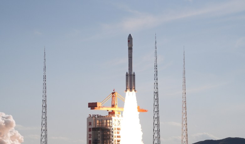 A Long March 6A rocket lifts off from the Taiyuan Satellite Launch Center in northern China, producing a bright plume of exhaust against a clear sky, with four lightning towers surrounding the launch pad.