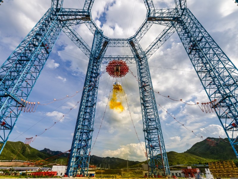 China’s Lanyue crewed lunar lander undergoing a landing and ascent test at the extraterrestrial celestial body landing test facility in Huailai, Hebei Province, on August 6, 2025. The lander is suspended from a large steel tower structure and firing its thrusters, producing a yellow exhaust plume characteristic of hypergolic propellants.
