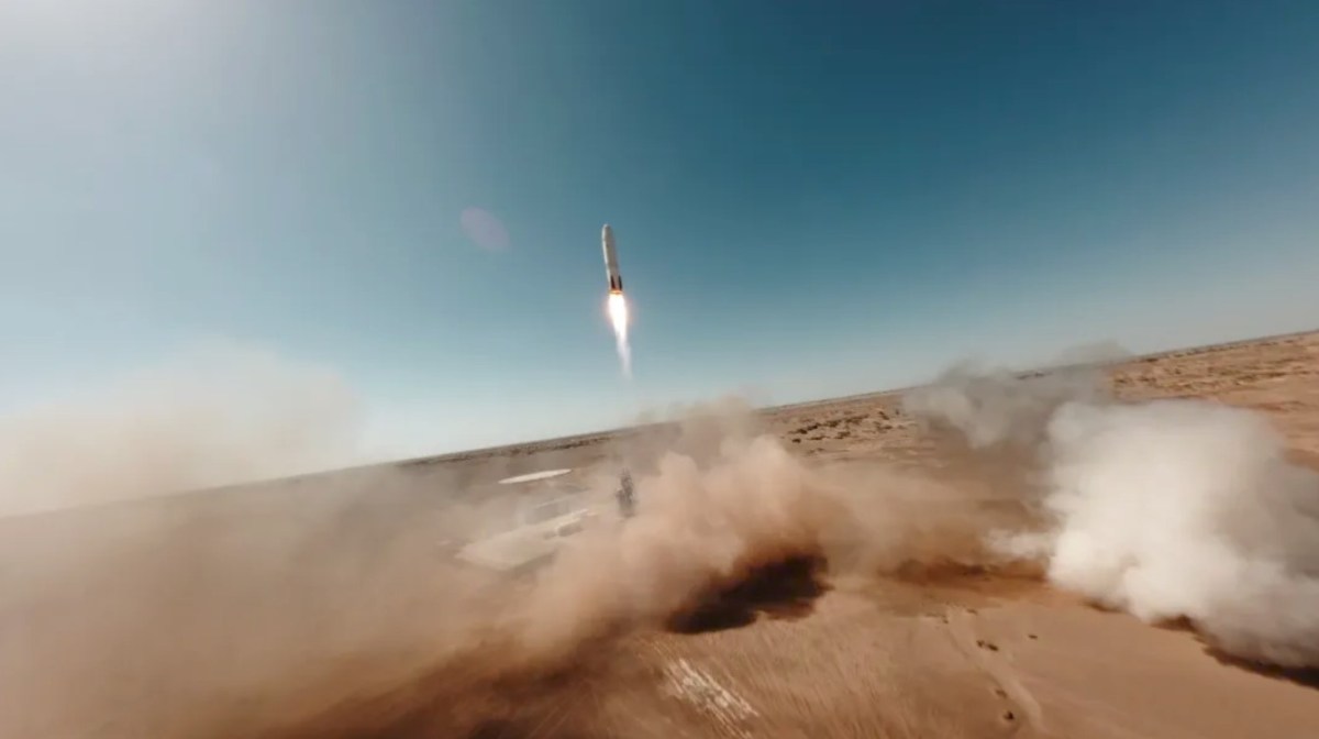Sand blows off a desert landing pad as a Nebula-1 first stage makes a powered descent from clear blue skies towards a landing pad.