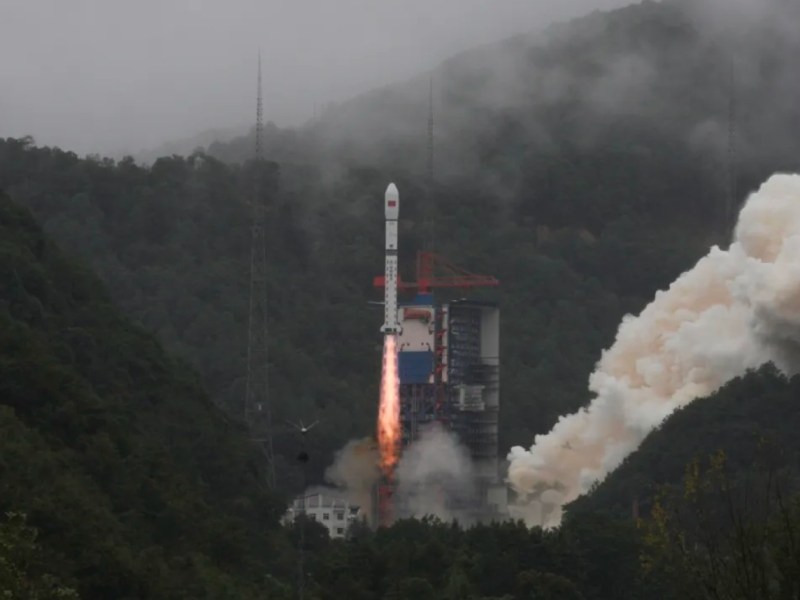 A Long March 2D rocket lifts off from the Xichang Satellite Launch Center in China, leaving a trail of flame and smoke against mist-covered hills.
