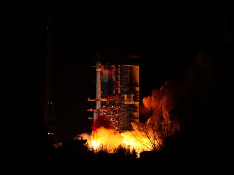 A Long March 3B rocket lifts off at night from the Xichang Satellite Launch Center, illuminating the surroundings with bright flames and smoke. The rocket is positioned on the launch pad with its structure and scaffolding visible, silhouetted against the dark sky.